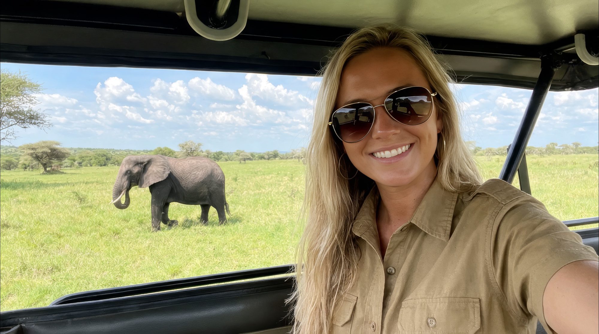 Mujer sonriente haciéndose un selfie en un vehículo de safari, detrás un elefante en la sabana