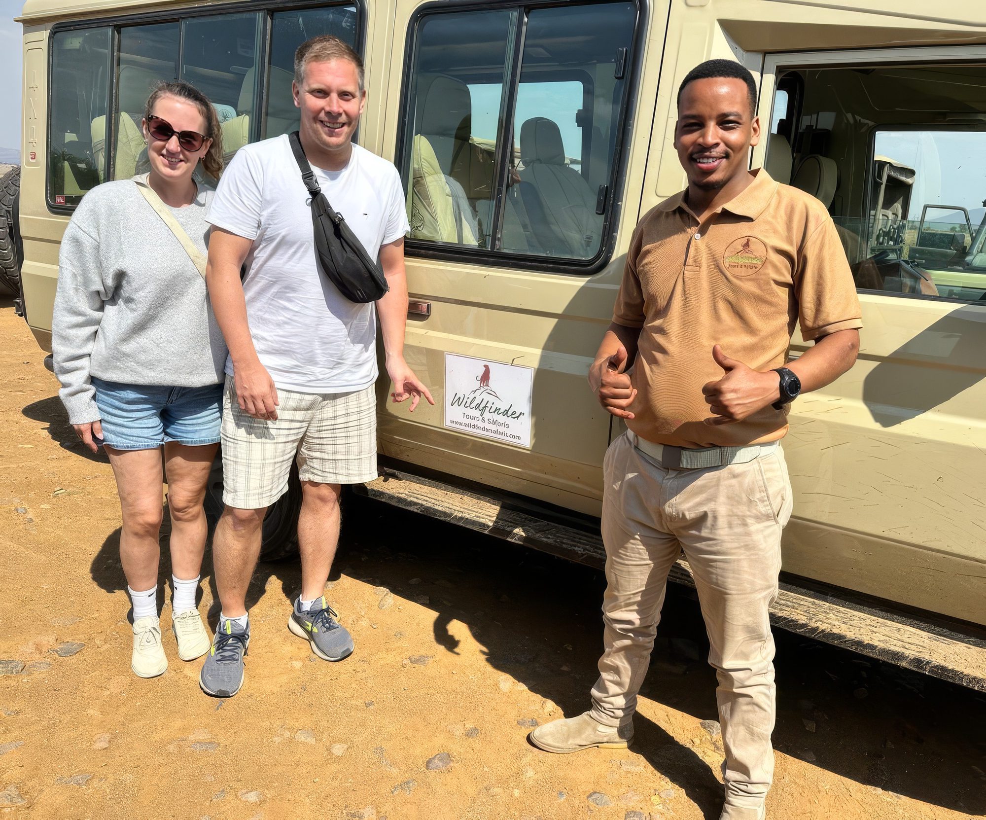 Zwei Gäste und ein Guide mit erhobenem Daumen vor einem Safarifahrzeug