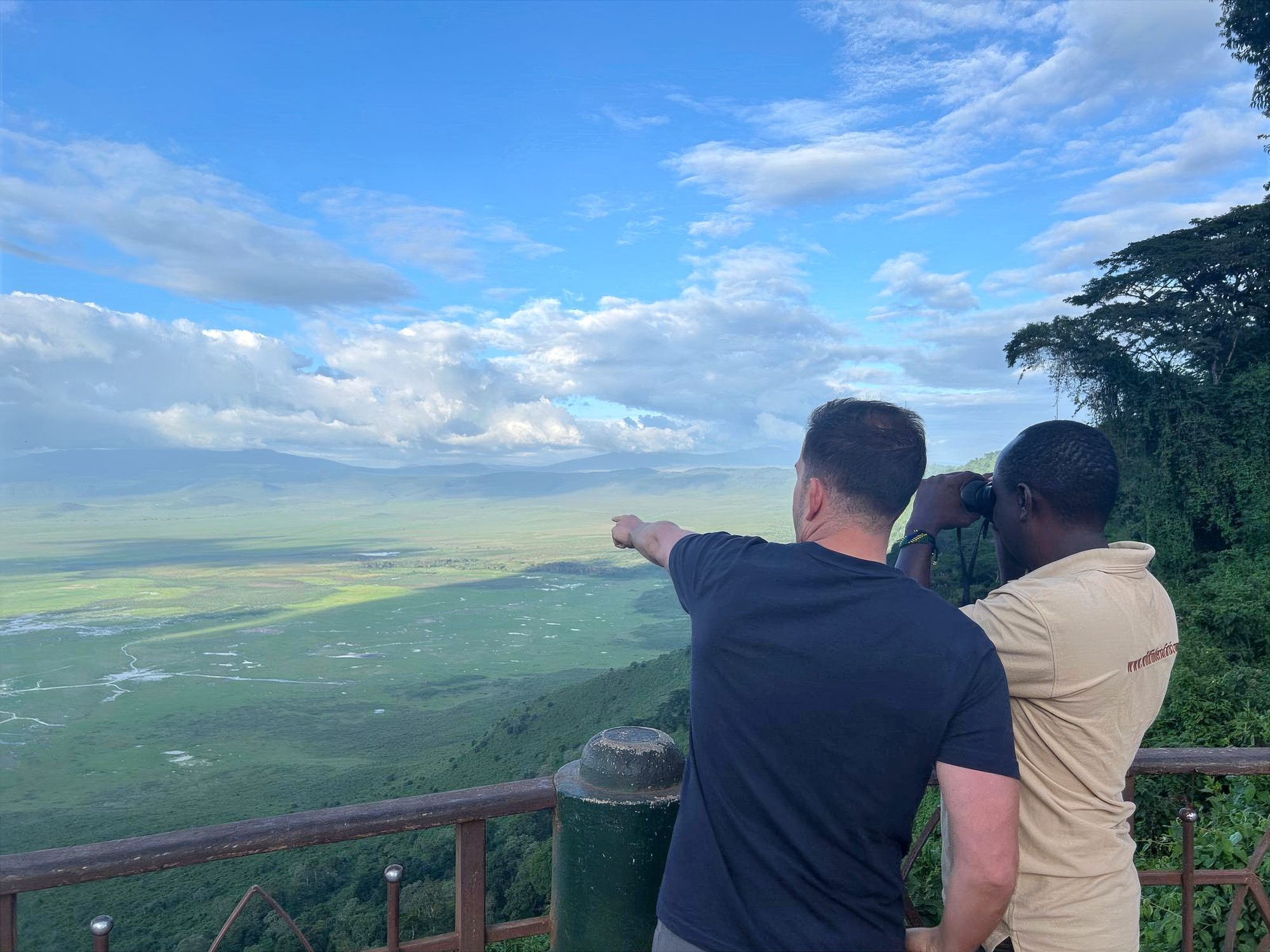 Zwei Männer am Holzgeländer über dem grünen Ngorongoro-Krater, einer zeigt hinein, einer schaut durchs Fernglas
