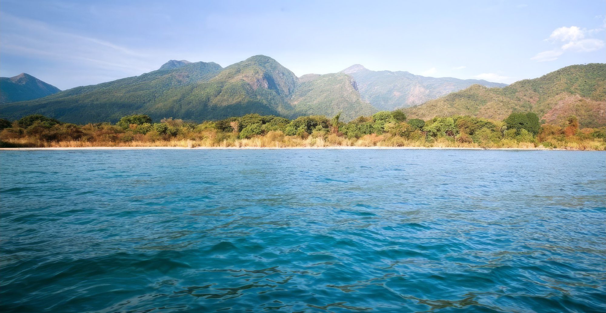 Türkisblaues Seewasser mit Wellen vor hellem Sandufer und steilen bewaldeten Bergen im Mahale-Mountains-Nationalpark