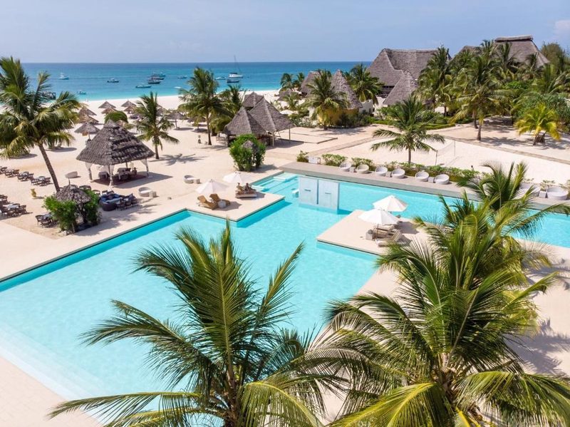 Pool mit Palmen und Strandhäusern im Hintergrund aus der Luft