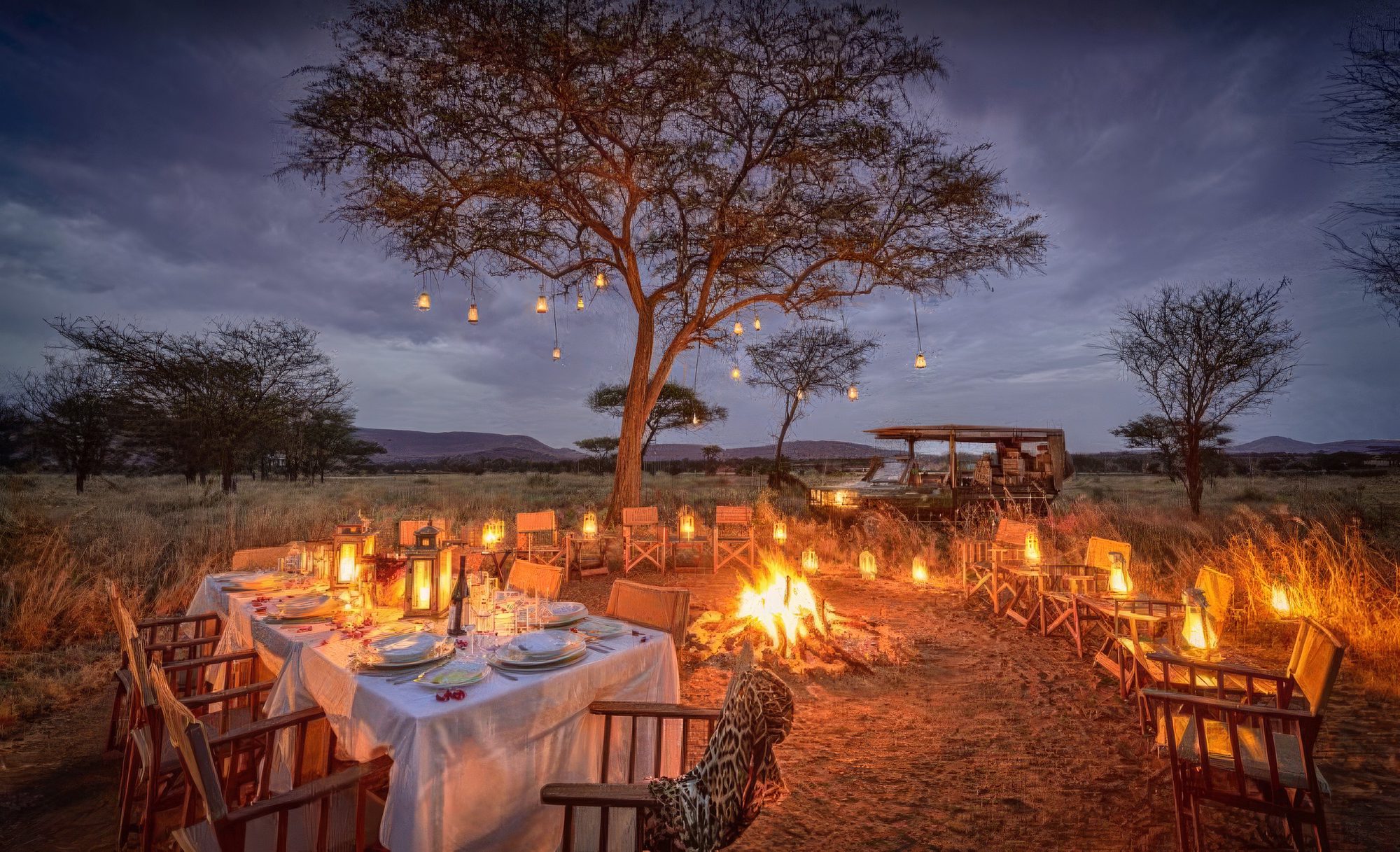 Hoguera y faroles en una acacia de Nimali Serengeti con mesa puesta y sillas plegables de safari