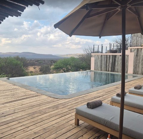 Infinity pool with wooden deck, loungers with rolled towels and a reed fence in front of bushland and hills
