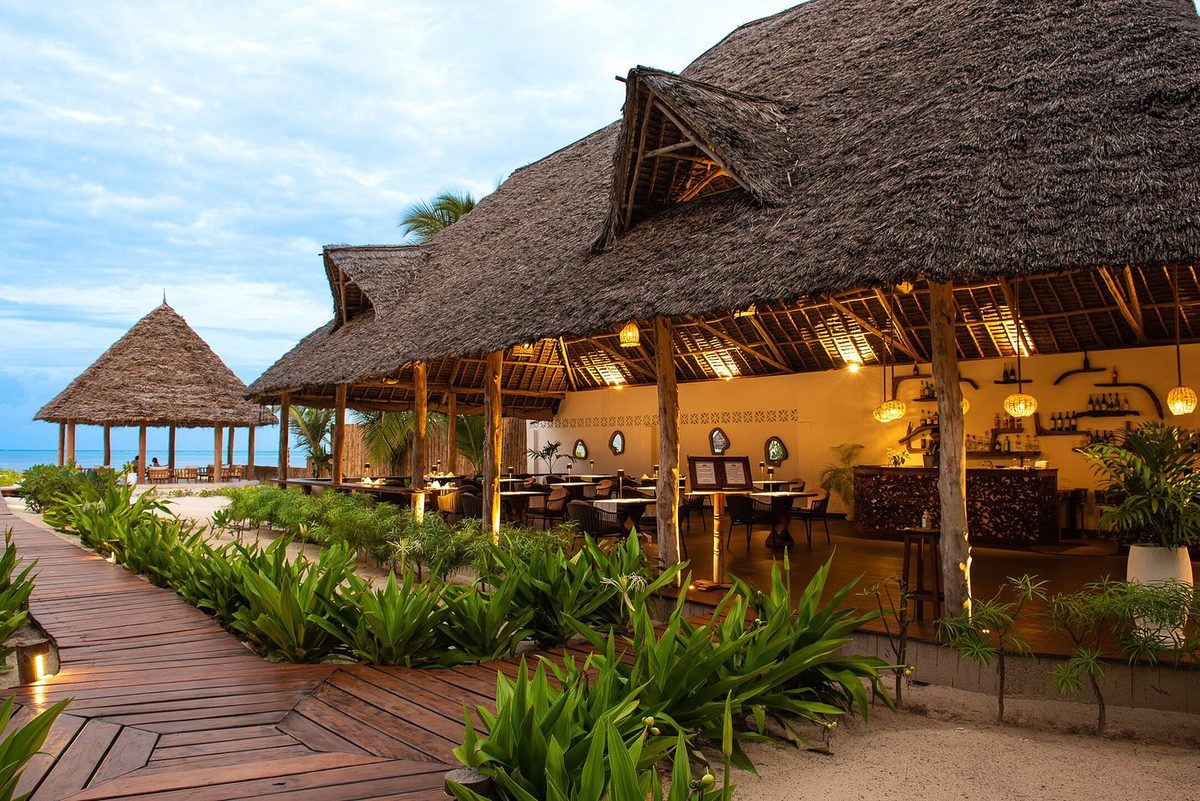 Restaurante del Amani Boutique Hotel bajo techo de paja con pasarela de madera y plantas por la noche