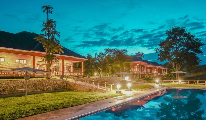 Helles Steinhaus von Baghayo Garden Suites mit roten Balken und Pfeilern, beleuchtetem Pool und Palmen in der Abenddämmerung