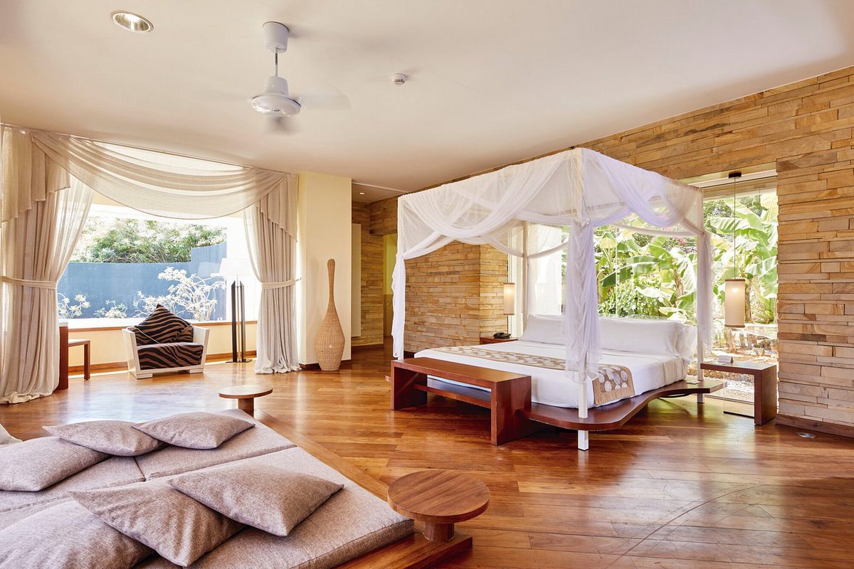 Habitación en el Hotel Riu Palace Zanzibar con cama con dosel, suelo de madera, pared de piedra natural y cojines de asiento