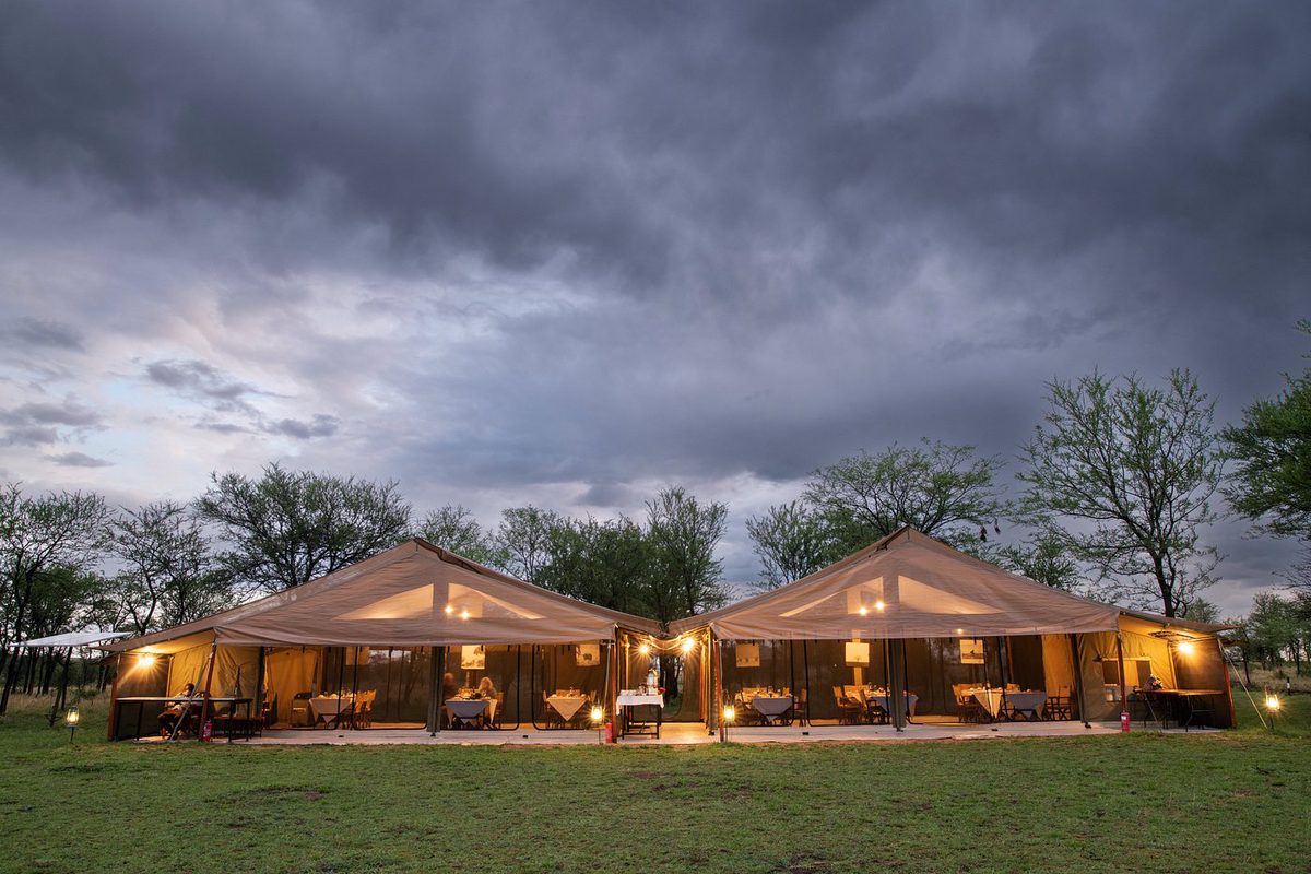 Tienda comedor iluminada del Ndutu Kati Kati Tented Camp con mesas puestas bajo un cielo oscuro de tormenta