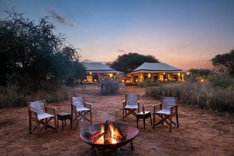 Hoguera con sillas frente a las tiendas iluminadas del Nyikani Camp Tarangire al anochecer