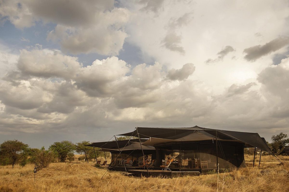 Tienda principal oscura del Siringit Migration Camp con zona de estar en la pradera seca bajo un cielo nublado
