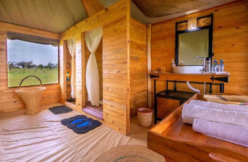 Zona de baño en una tienda de Tanzania Bush Camps con paredes de madera, lavabo y ventana a la pradera