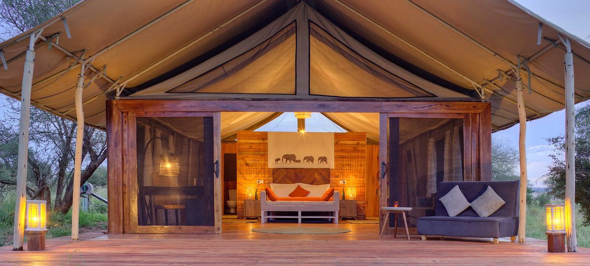 Tienda iluminada de la Tarangire Ndovu Tented Lodge con frente abierto, cama y sofá en la terraza de madera