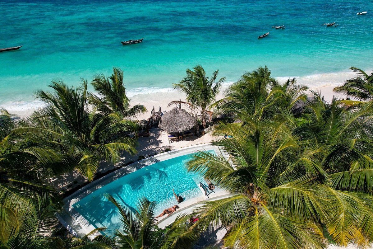 Vista aérea de la piscina de la Villa Juliana bajo palmeras en la playa con mar turquesa y barcas