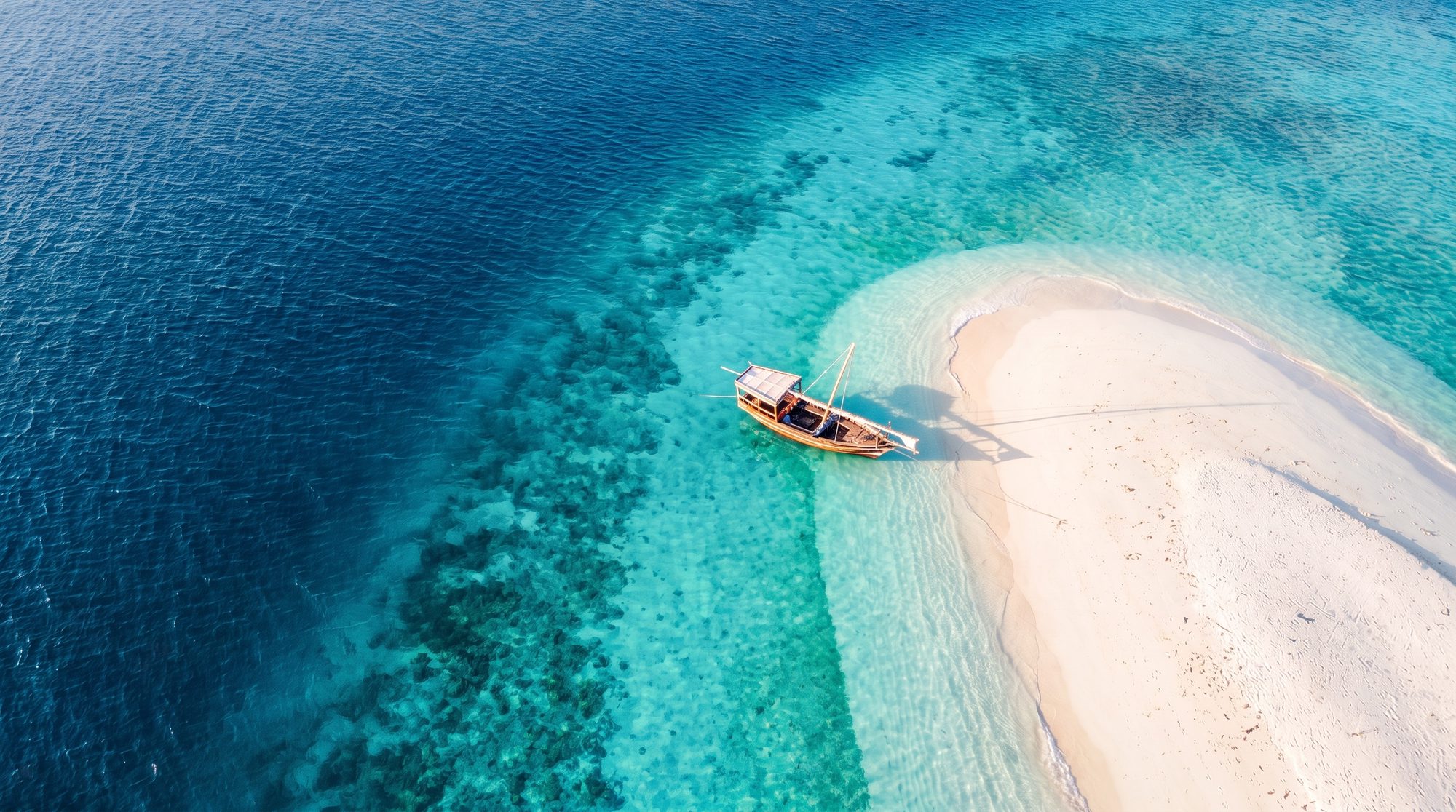 Aerial view of a wooden dhow beside a white sandbank between turquoise shallows and deep blue sea