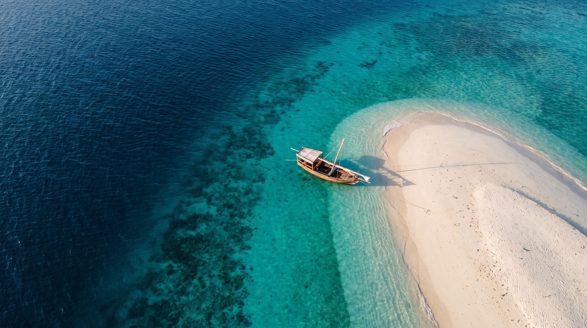 Luftaufnahme: hölzerne Dhow an weißer Sandbank zwischen türkisfarbenem Flachwasser und tiefblauem Meer