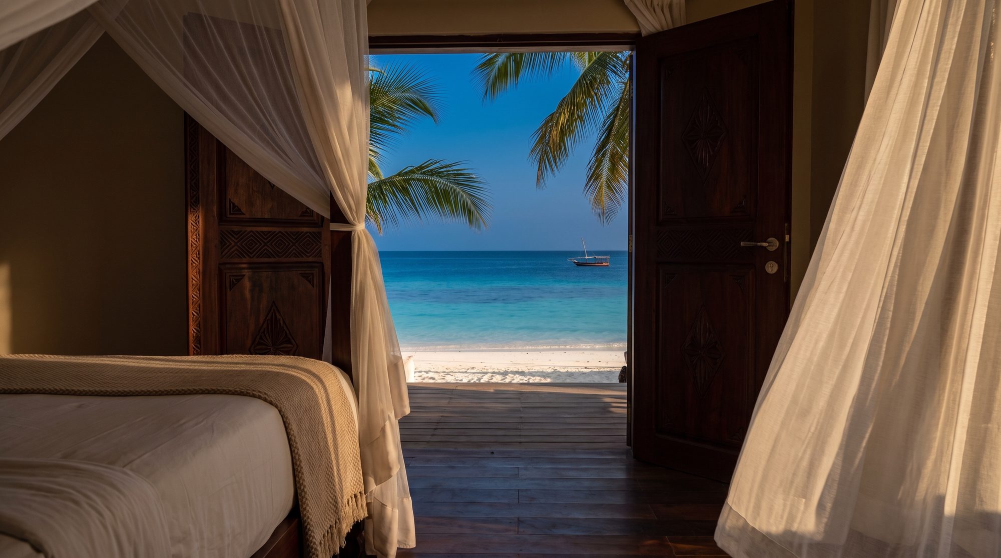 Himmelbett mit Moskitonetz, offene geschnitzte Holztür, dahinter Terrasse, weißer Strand und Meer mit Boot