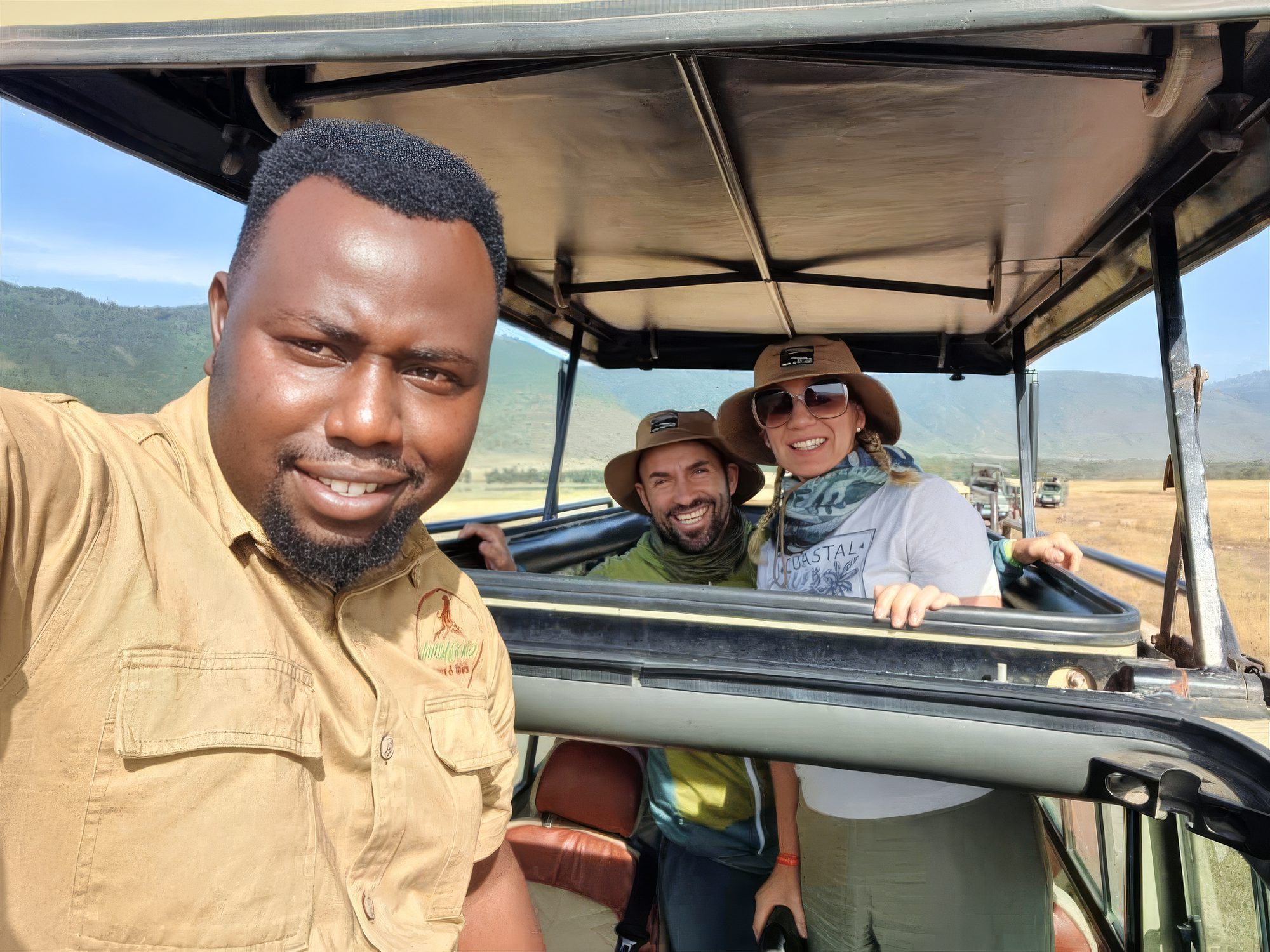 Selfie by a guide, two guests wearing safari hats standing and smiling in the roof hatch of a safari vehicle